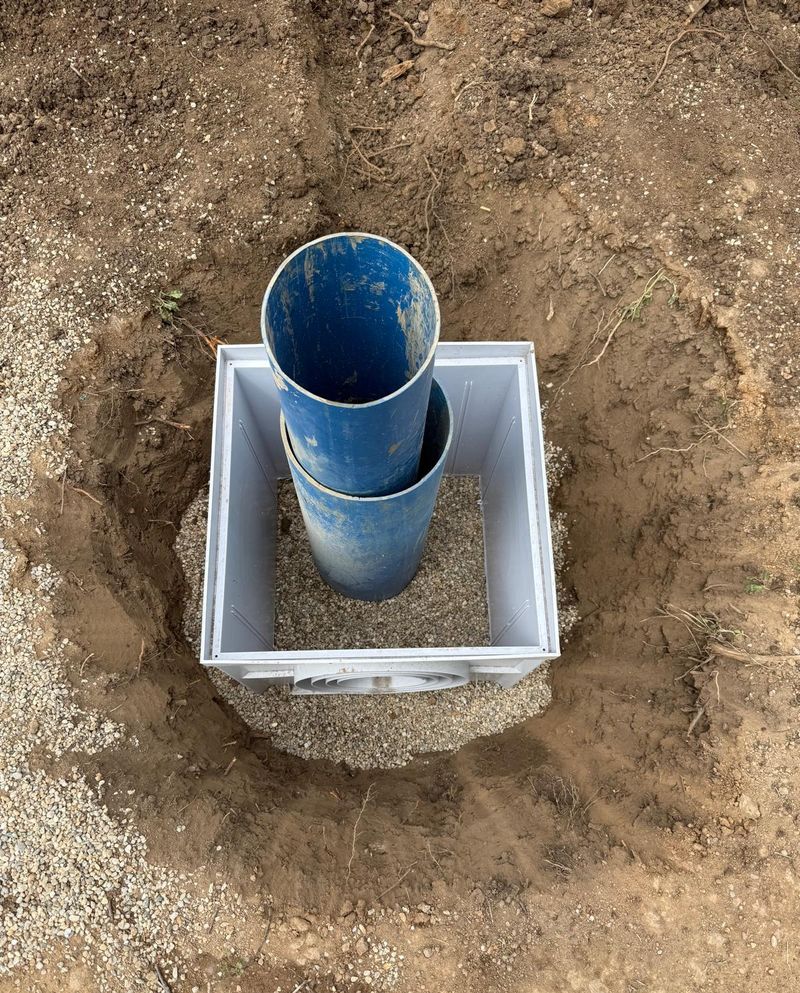 Cămin de vizitare din plastic gri, așezat pe un pat de pietriș în groapă, în jurul coloanei albastre de PVC a puțului, văzut de sus