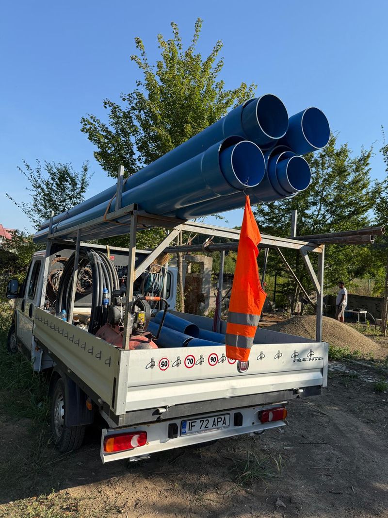 Camioneta firmei văzută din spate, cu tuburi albastre de PVC pe rastel, compresorul și sculele în benă, lângă o grămadă de pietriș