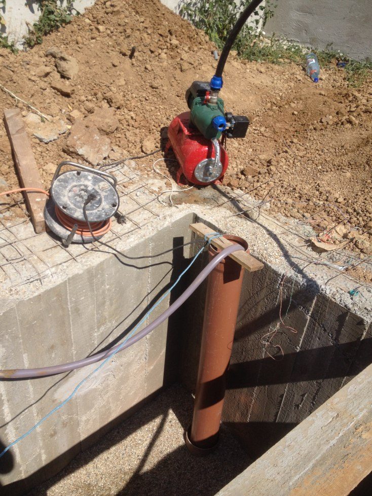 Hidrofor de suprafață cu motor verde și vas roșu, așezat pe pământul săpat lângă o groapă de beton în care intră un tub portocaliu; alături, un tambur cu cablu
