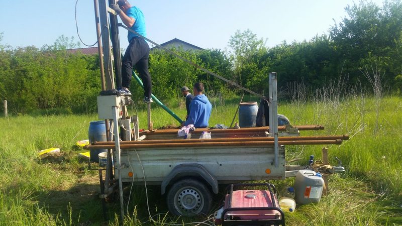 Instalație de foraj pe remorcă, cu tije și generator, la marginea unui teren cu casă în fundal, fotografie din aprilie 2018