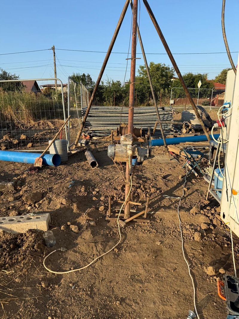 Instalație de foraj cu trepied, motor electric și troliu, pe terenul unei case noi, cu tuburi albastre de PVC pe jos
