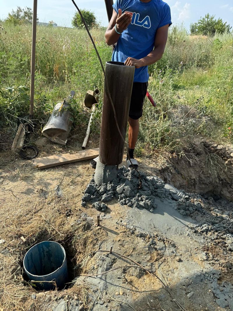 Lingura de foraj din oțel, ridicată din puț, cu nisip cenușiu ud la bază, lângă coloana albastră de PVC dintr-o groapă