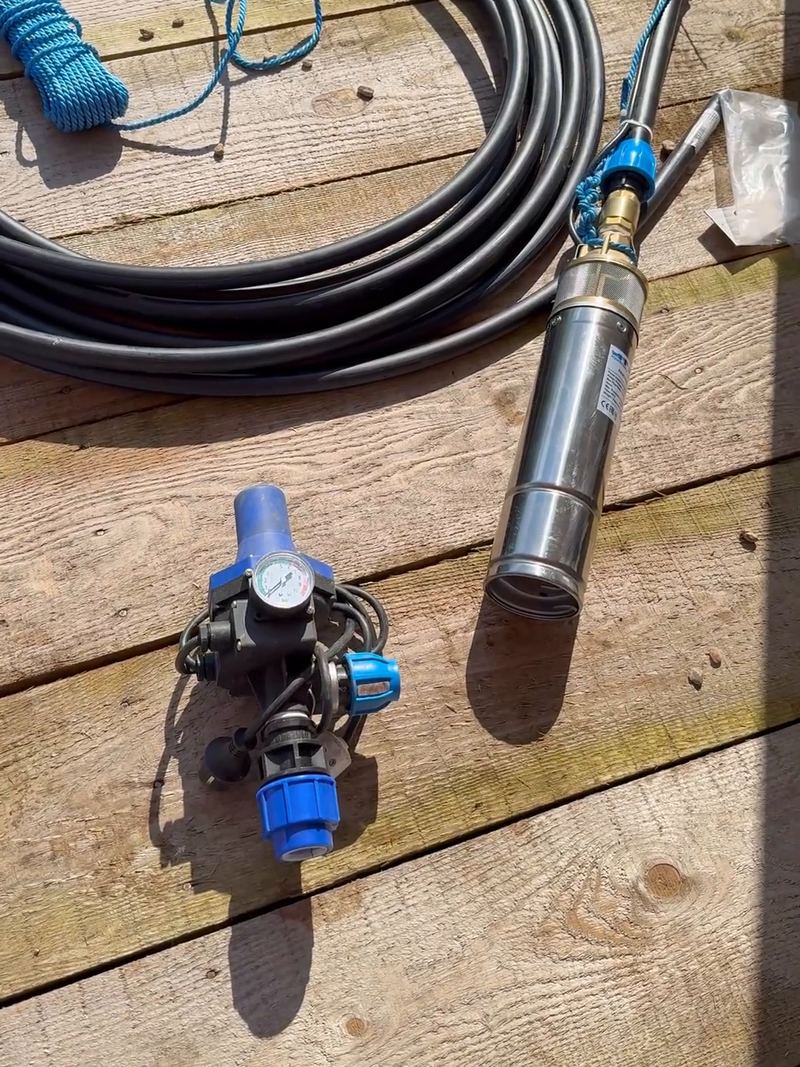 Pompa submersibilă din inox, automatizarea cu manometru și racordurile albastre, lângă un colac de conductă neagră de polietilenă, pe o terasă de lemn, înainte de montaj, în Balotești