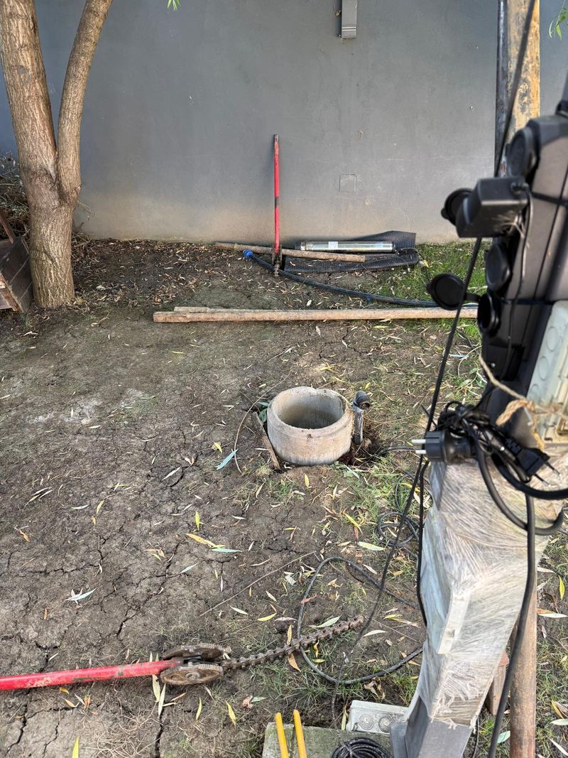 Puț din tuburi de beton în mijlocul curții; lângă zid, pompa submersibilă din inox scoasă din puț, pe o folie, și tijele de foraj