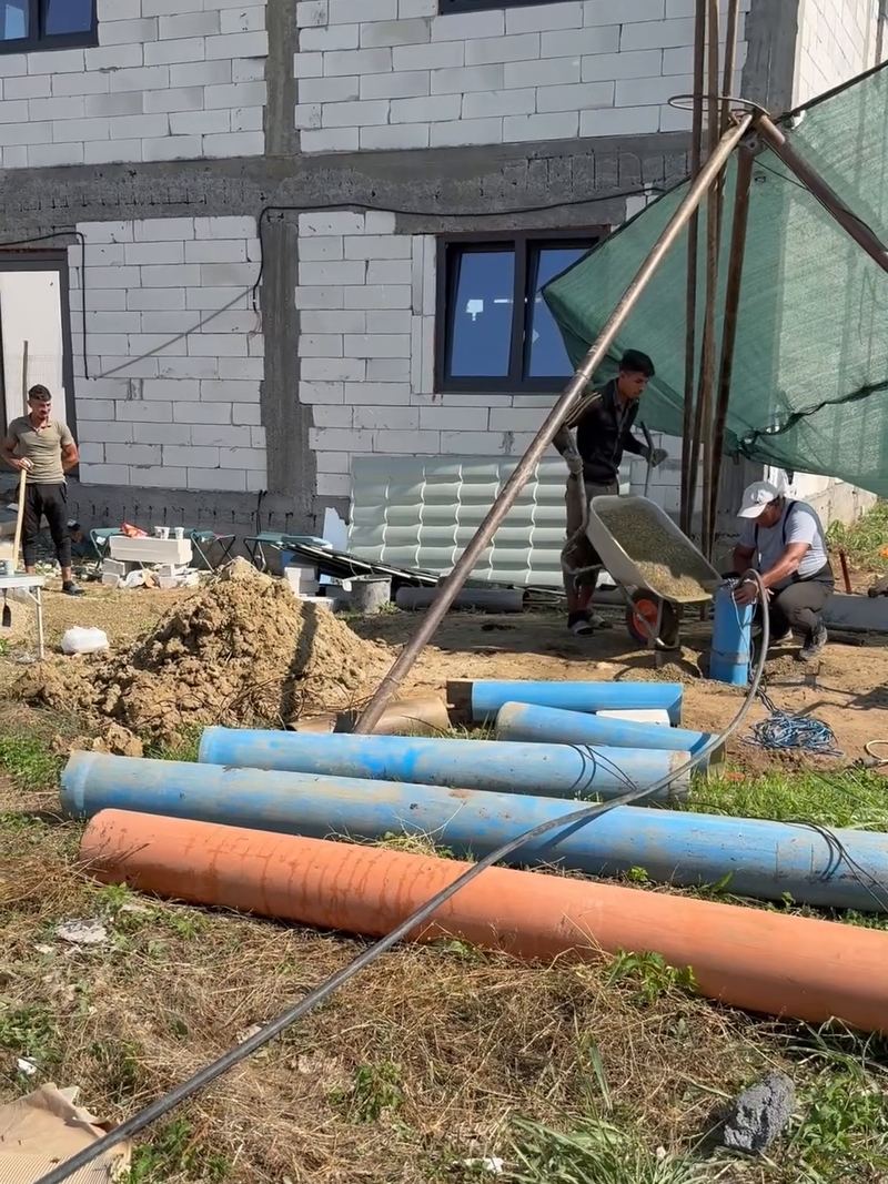 Sonda de foraj a firmei, cu prelată verde, lângă o casă nouă în construcție; în față, tuburile de PVC albastre și portocalii pentru coloana puțului, Buftea