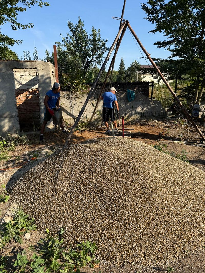Grămadă de pietriș mărgăritar în prim-plan și, în spate, trepiedul de foraj cu doi oameni din echipă la lucru