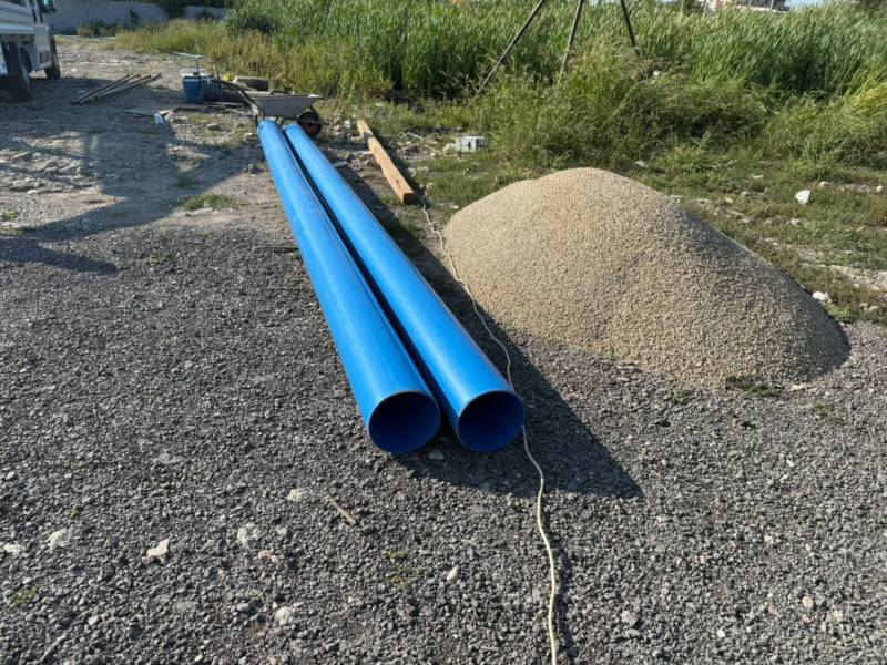 Două tuburi groase de PVC albastru pentru tubarea unui puț, întinse lângă o grămadă de pietriș mărgăritar pentru filtru, cu trepiedul de foraj în spate