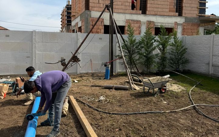 Trepied de foraj manual cu troliu, într-o curte împrejmuită de casă nouă, cu un muncitor care lucrează la tubul albastru de PVC al puțului, 2026